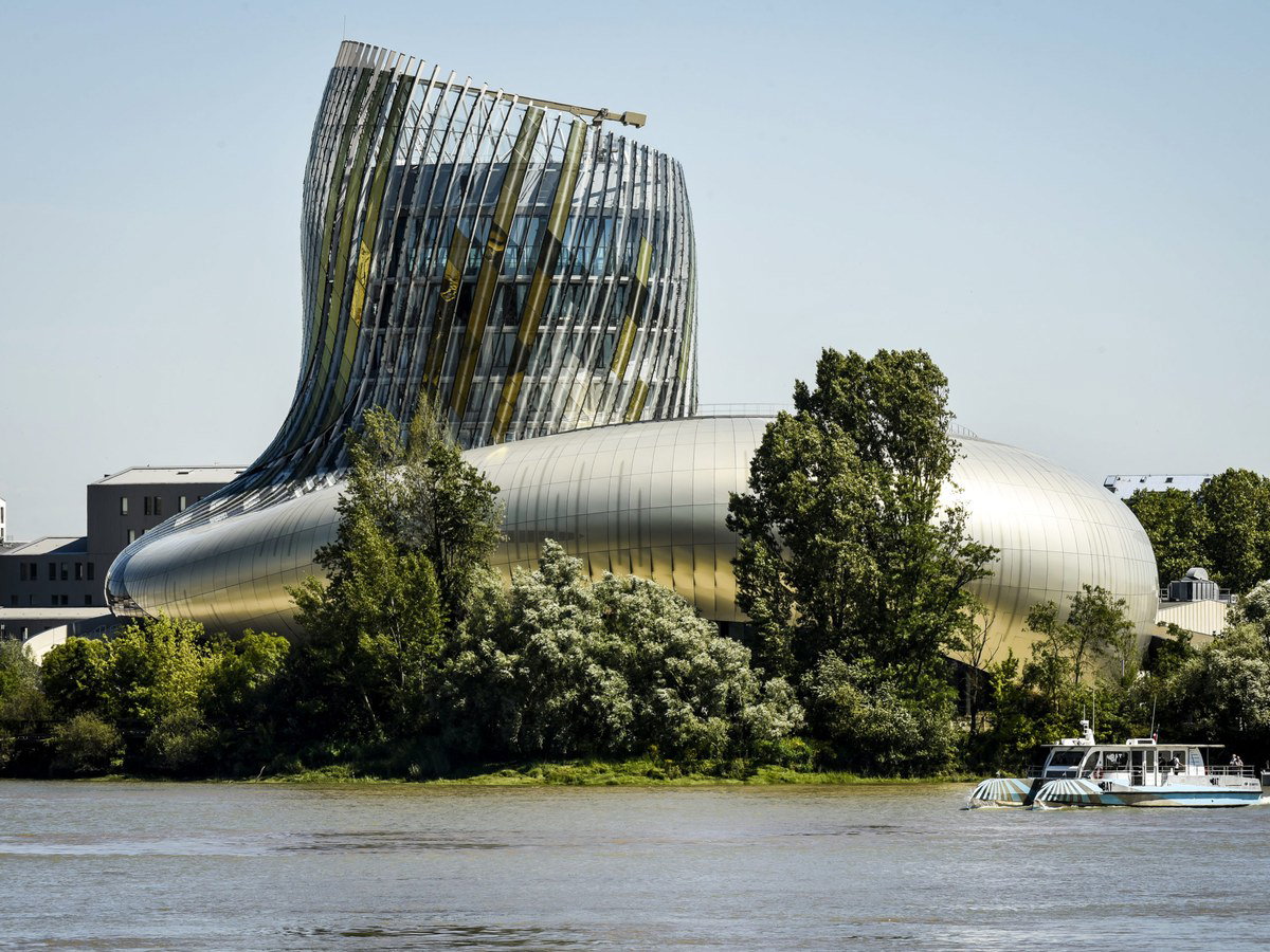 The Cité du Vin building in Bordeaux, with its distinctive undulating gold and silver metallic facade, rises from a rounded base with a glass and