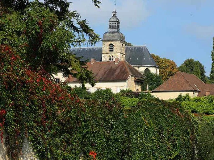 A stone church with a prominent bell tower rises above buildings with red-tiled roofs, partially obscured by a lush green and red ivy-covered wall.