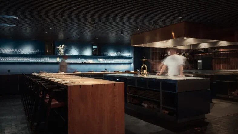 A dark, moody open-kitchen bar/lounge interior with a long wooden counter, blue-tiled back wall, and motion-blurred staff.