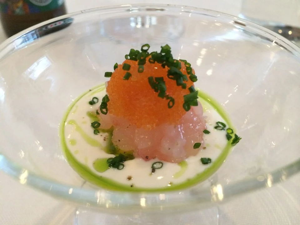 A delicate dish of raw seafood topped with bright orange roe and chopped chives, served in a clear glass bowl with a white cream sauce and green herb