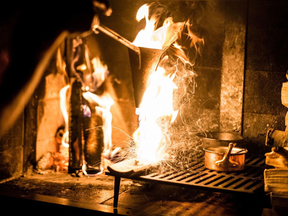 A cook pours flaming liquid from a conical vessel over a wood-burning hearth, with sparks flying and copper pots on a grate.