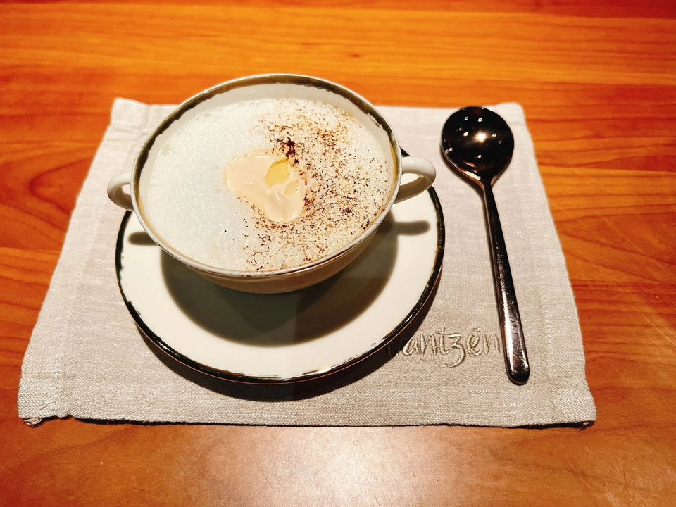 A white cup with a gold rim, filled with a foamy beverage and topped with cocoa powder, sits on a saucer and a linen napkin embroidered with