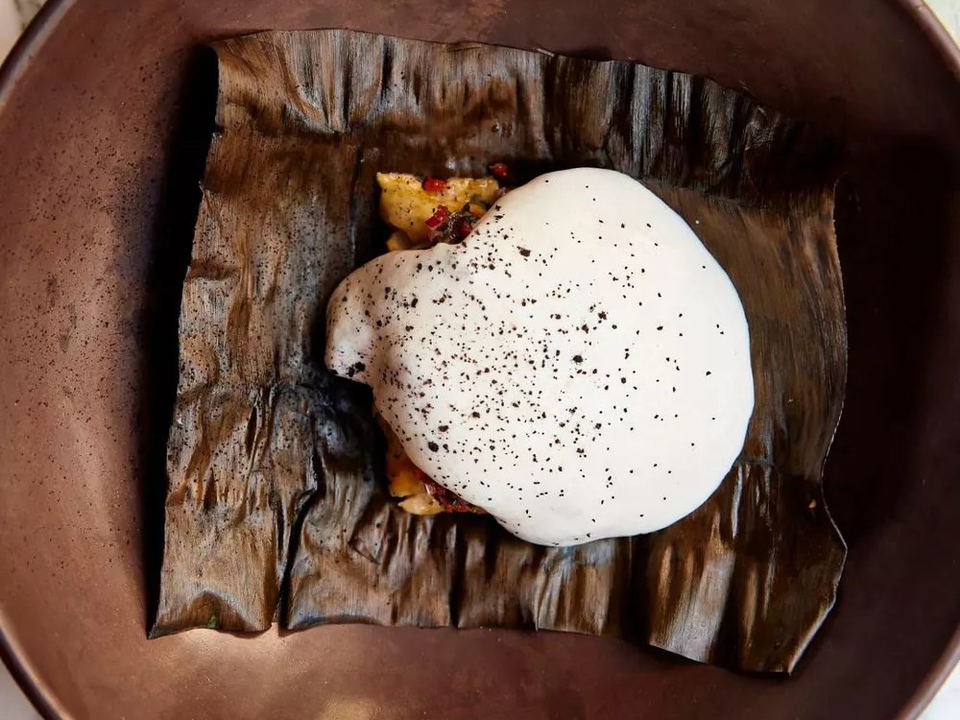 An elegantly plated tasting-menu course, featuring a mound of filling on a charred banana leaf topped with a white foam dusted with black specks,