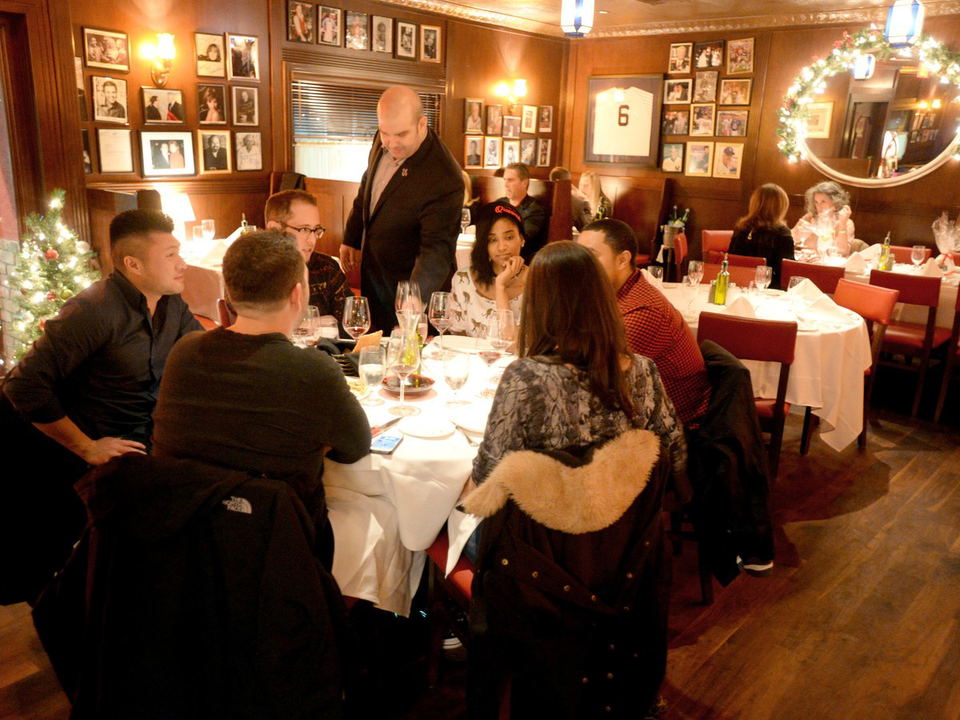 A warmly lit dining room at Rao's, with wood-paneled walls adorned with framed celebrity photos, red booths, and a group of diners at a table.