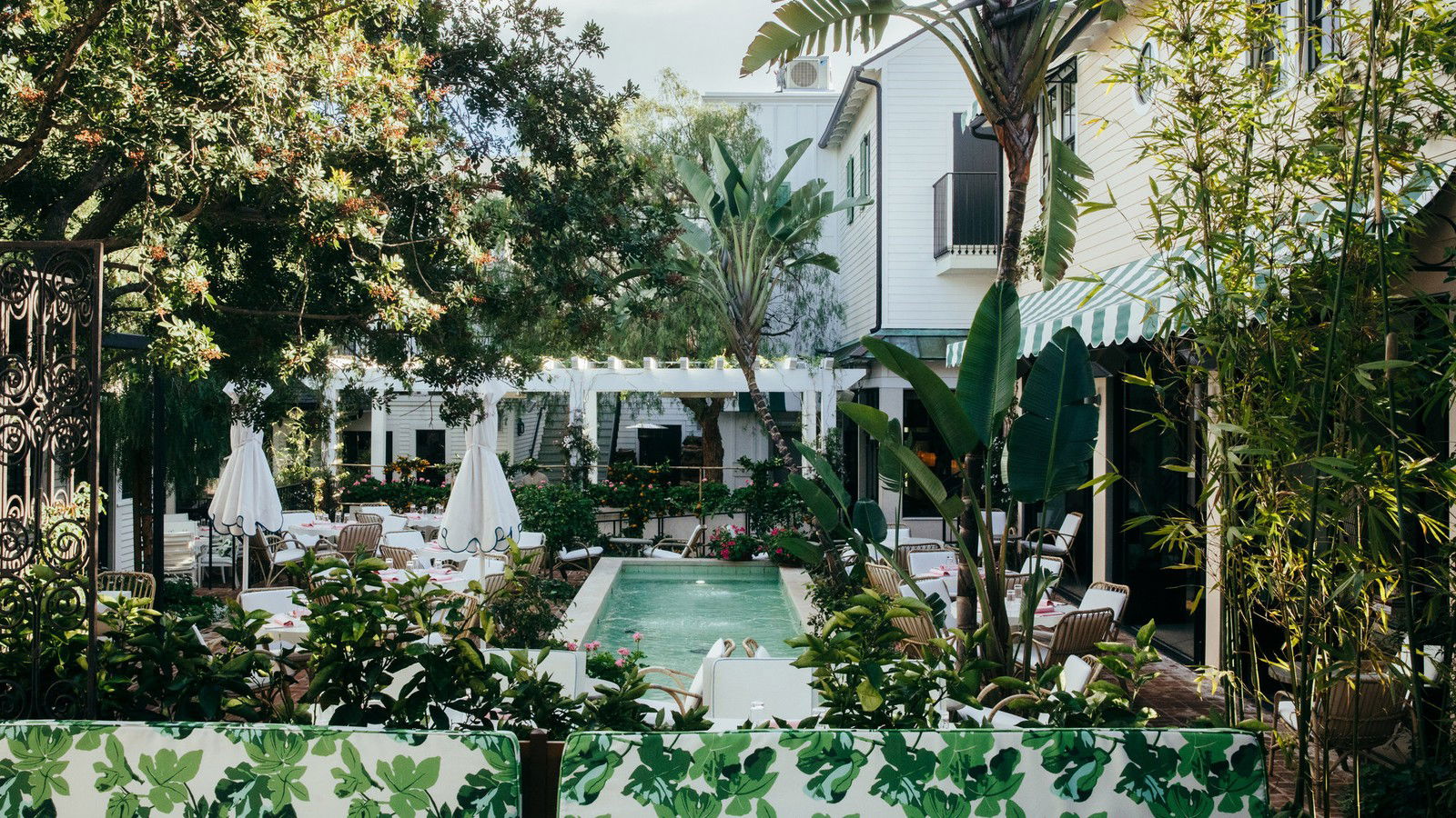 An outdoor garden courtyard at San Vicente Bungalows features a narrow pool, lush tropical plants, white bungalows, and dining tables.
