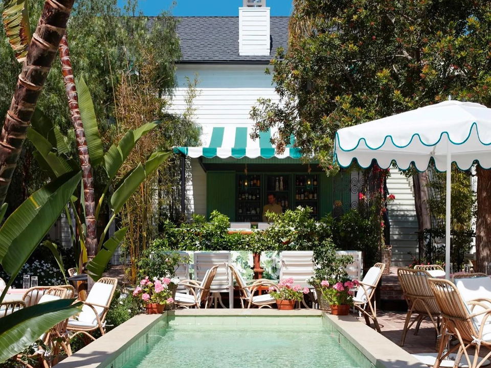 A narrow outdoor lap pool with turquoise tiles, surrounded by lush tropical plants, rattan chairs, and a white building with a green-striped awning.