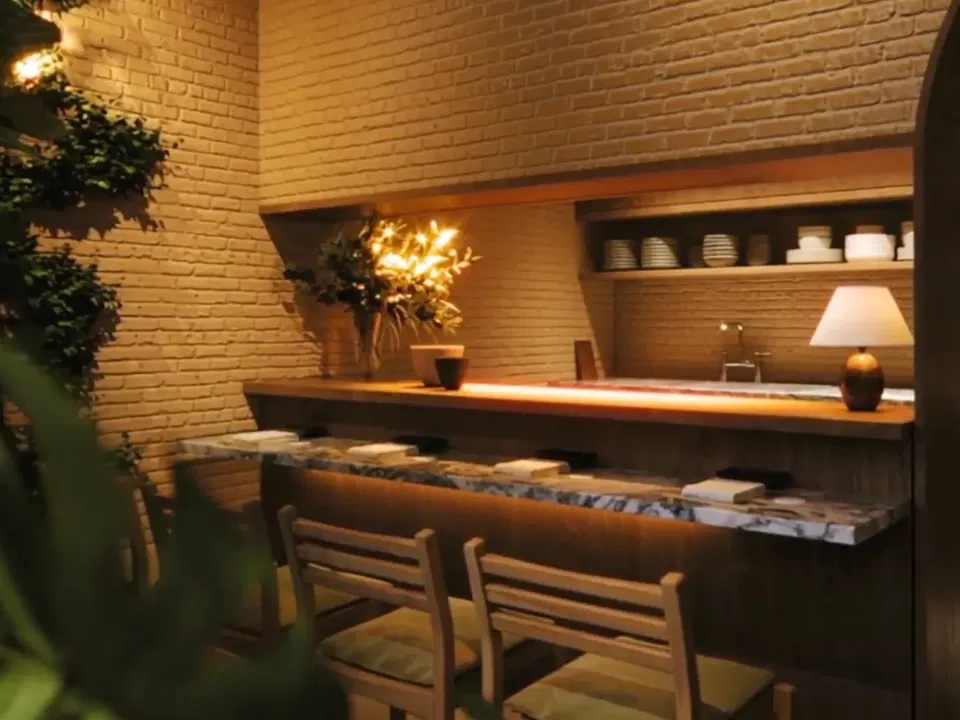 A modern reception area with brick and wood walls, a long desk with a table lamp, decorative plants, and open shelving with white dishware and linens.