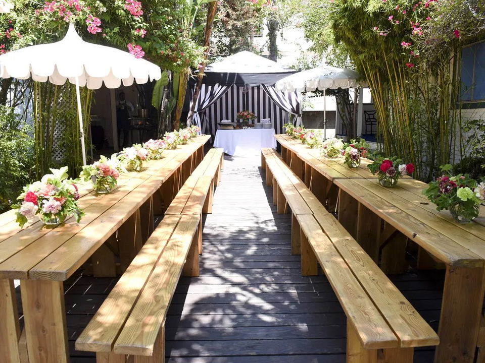 An outdoor dining area at San Vicente Bungalows features long wooden tables with benches, white umbrellas, and floral arrangements.