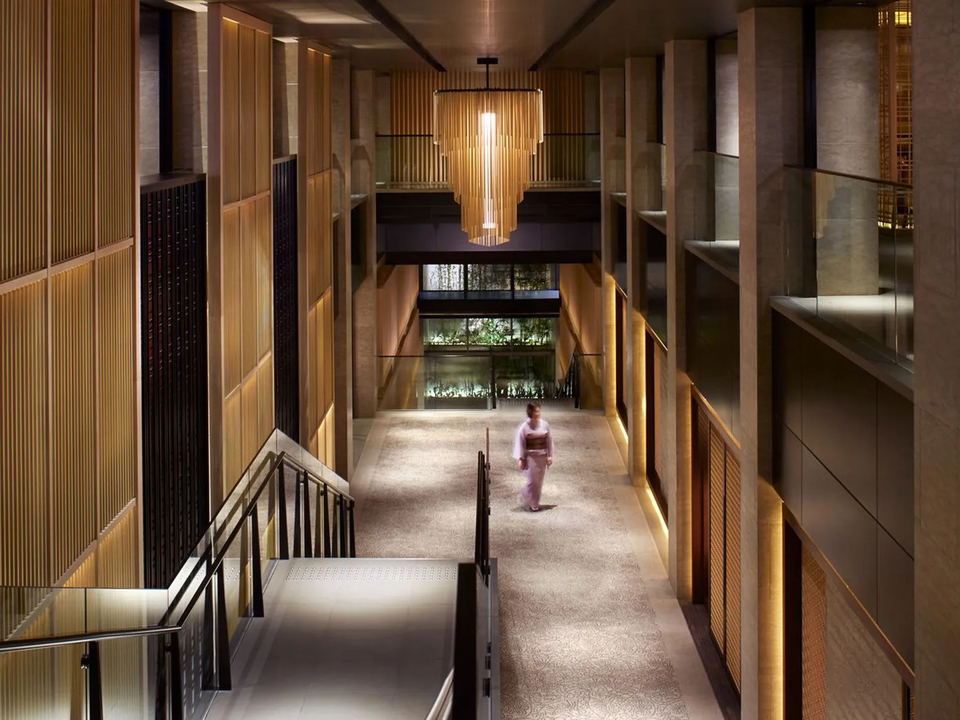 A multi-story hotel interior corridor with warm wood slat paneling, a dramatic pendant chandelier, and a figure in a kimono walking.