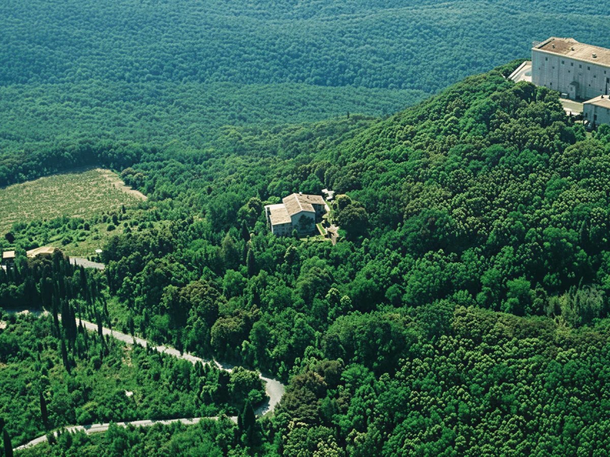 An aerial view shows a large, light-colored building with a flat roof atop a densely forested hill, with smaller structures and winding roads below.