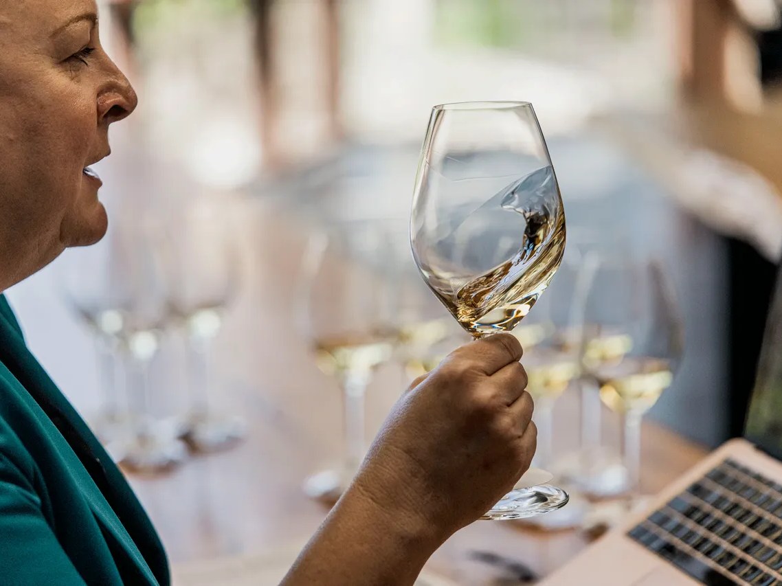 A woman in a teal top swirls white wine in a glass, with other glasses and a laptop visible in the blurred background.