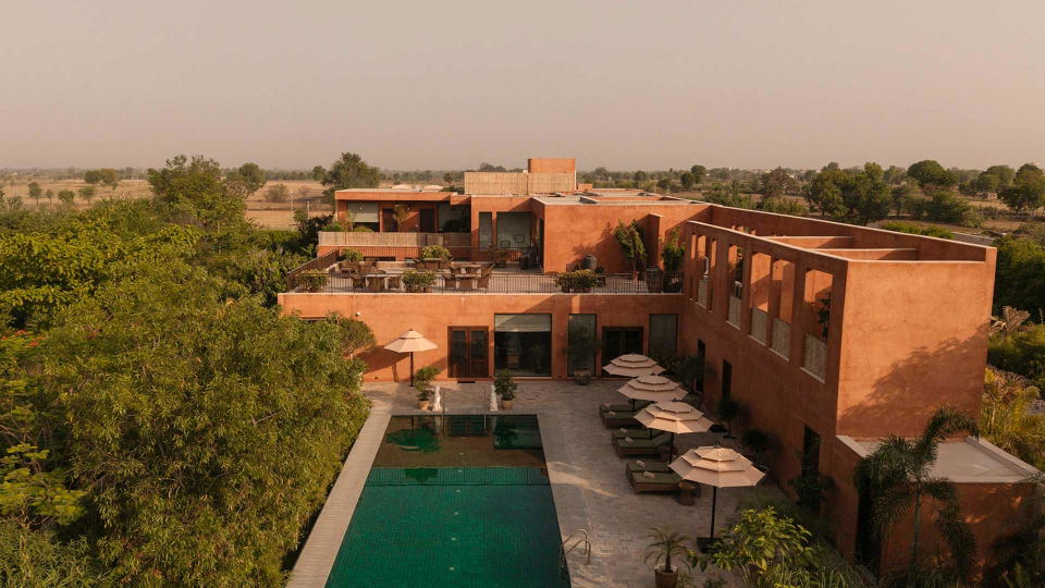 Aerial view of a contemporary orange-brick resort building with a green swimming pool, sun loungers with umbrellas, and surrounding vegetation and fields.