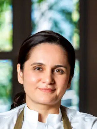 A professional portrait of Chef Garima Arora, wearing a white chef's coat and an olive apron, with her arms crossed.