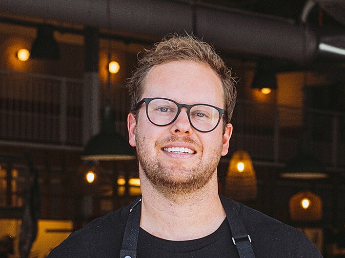 A smiling man wearing glasses and a black chef's apron over a black t-shirt stands in front of a restaurant bar interior.