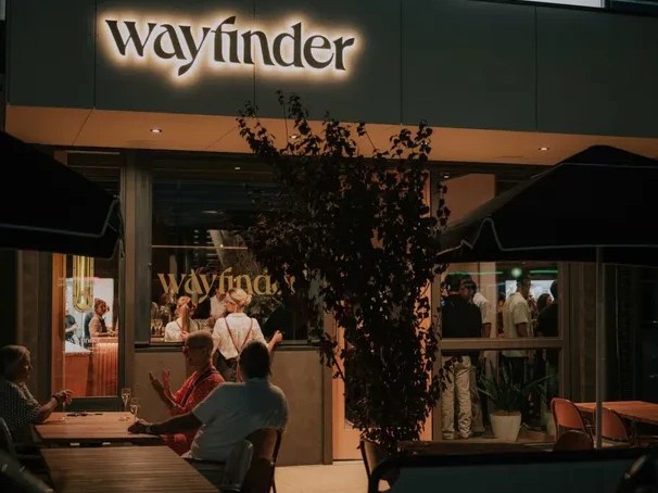 Nighttime exterior of Wayfinder Wines, showing the illuminated 'wayfinder' sign, outdoor seating with patrons, and people inside through glass