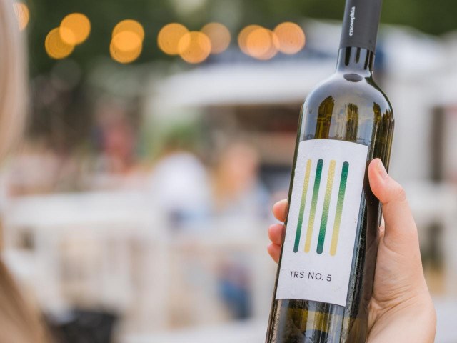 A hand holds a bottle of 'TRS NO. 5' wine from Enosophia Winery against a blurred outdoor background with bokeh lights.