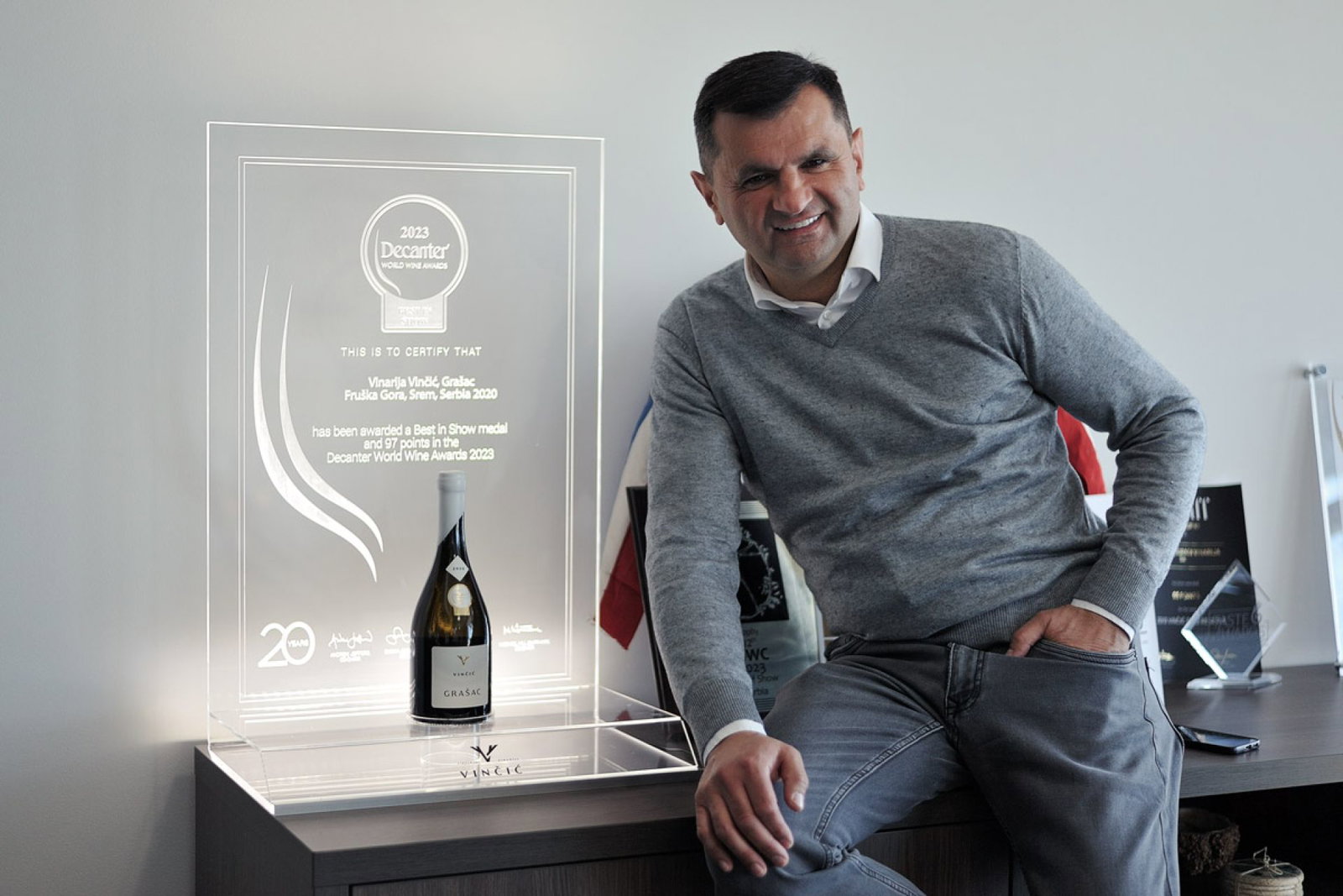 A man in a grey sweater smiles while sitting next to a clear acrylic award displaying a bottle of Vinarija Vinčić Grašac wine.