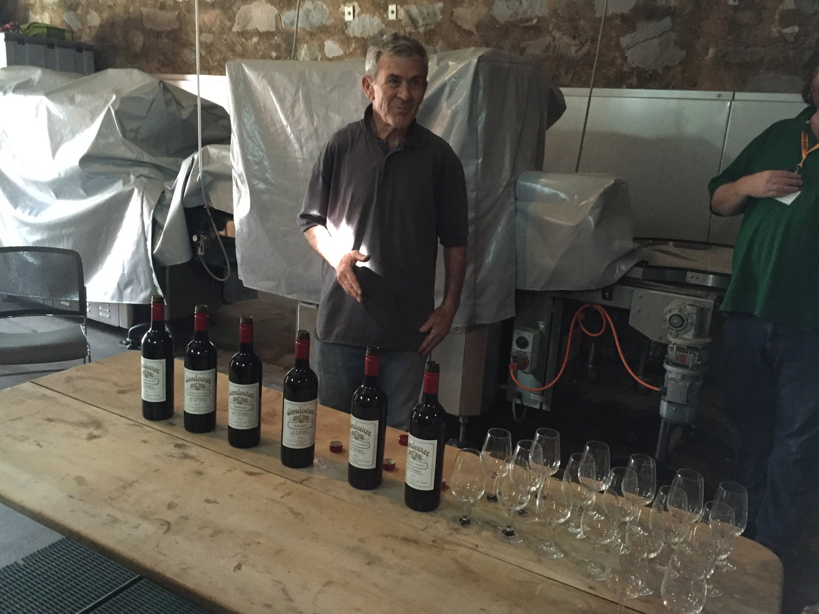 An older man in a dark polo shirt stands behind a wooden table lined with six Wendouree red wine bottles and rows of empty tasting glasses, inside a stone-walled winery cellar.