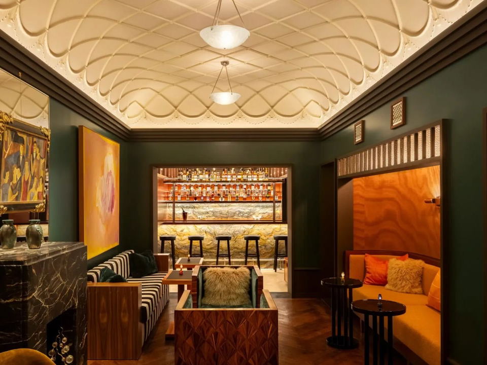 An elegant, intimate cocktail lounge with dark green walls, a patterned ceiling, a marble fireplace, and a backlit bar.