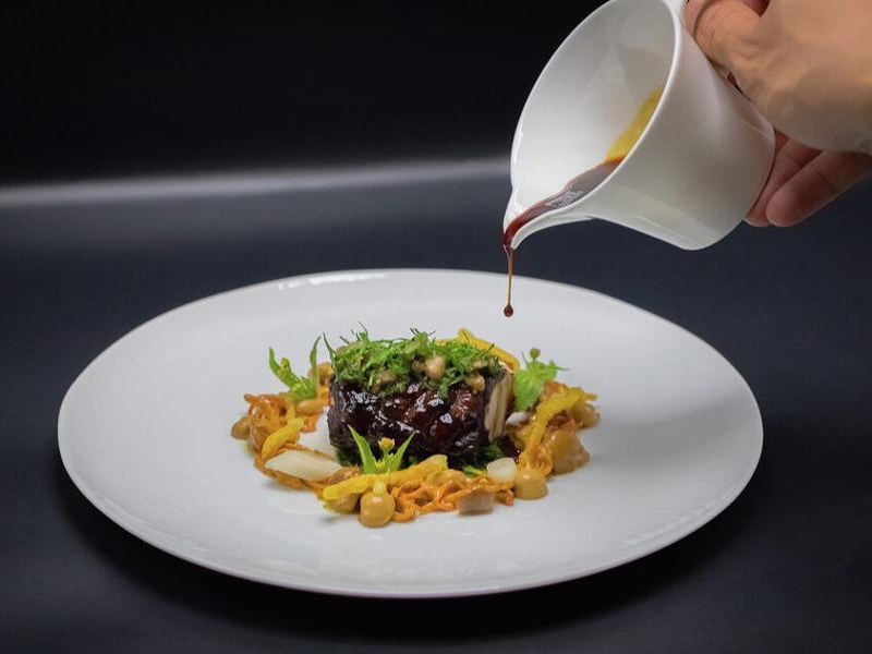 An elegantly plated fine-dining course on a white plate, featuring a glazed protein centerpiece garnished with microgreens, mushrooms, and golden.