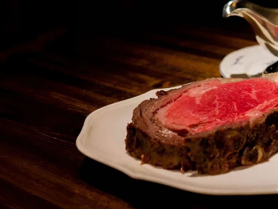 A thick slice of prime rib with a deeply seared exterior and a vivid red, rare interior is presented on a white plate, accompanied by a small ramekin