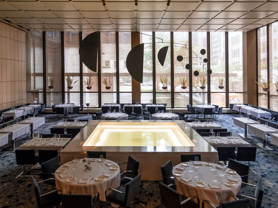 A spacious, modern dining room with floor-to-ceiling windows, white-clothed tables, and a central illuminated pool feature.