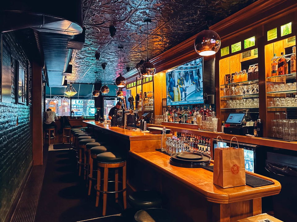 Corner Bistro East: A classic American bar with a long wooden counter, stools, tin ceiling, and a TV showing sports.