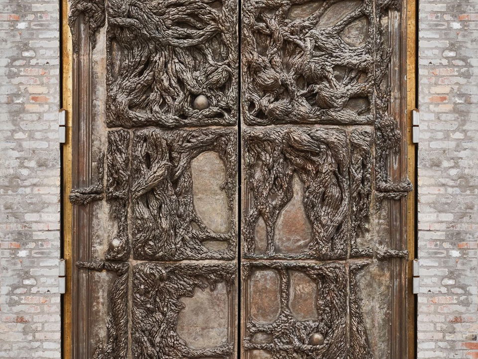 A pair of massive ornate bronze doors with intricate root and vine relief sculpture, set into a brick wall, marking the entrance to Alchemist.