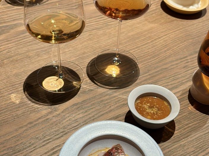 Three wine glasses containing golden-colored wine arranged on black coasters, with a white plate containing food and a small white bowl with sauce on a wooden table.