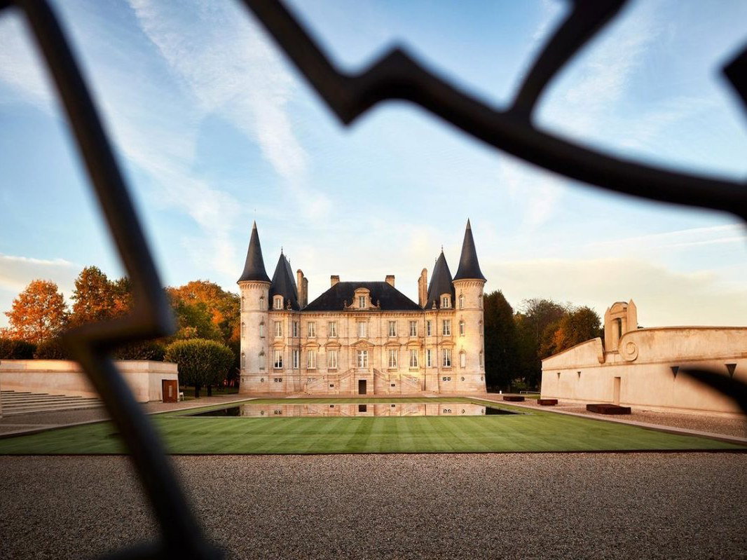 A grand French Château Pichon Baron with four conical-roofed towers, a manicured lawn, and a reflective pool, seen through an ornate iron gate.