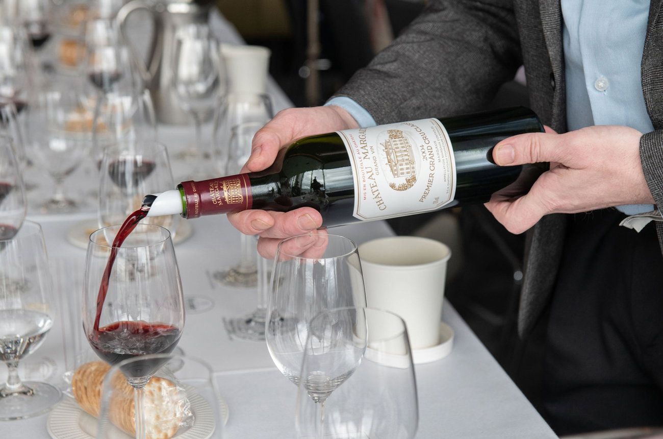 A person in business attire pours red wine from a bottle into a wine glass at a formal table setting with place settings and other glassware visible.