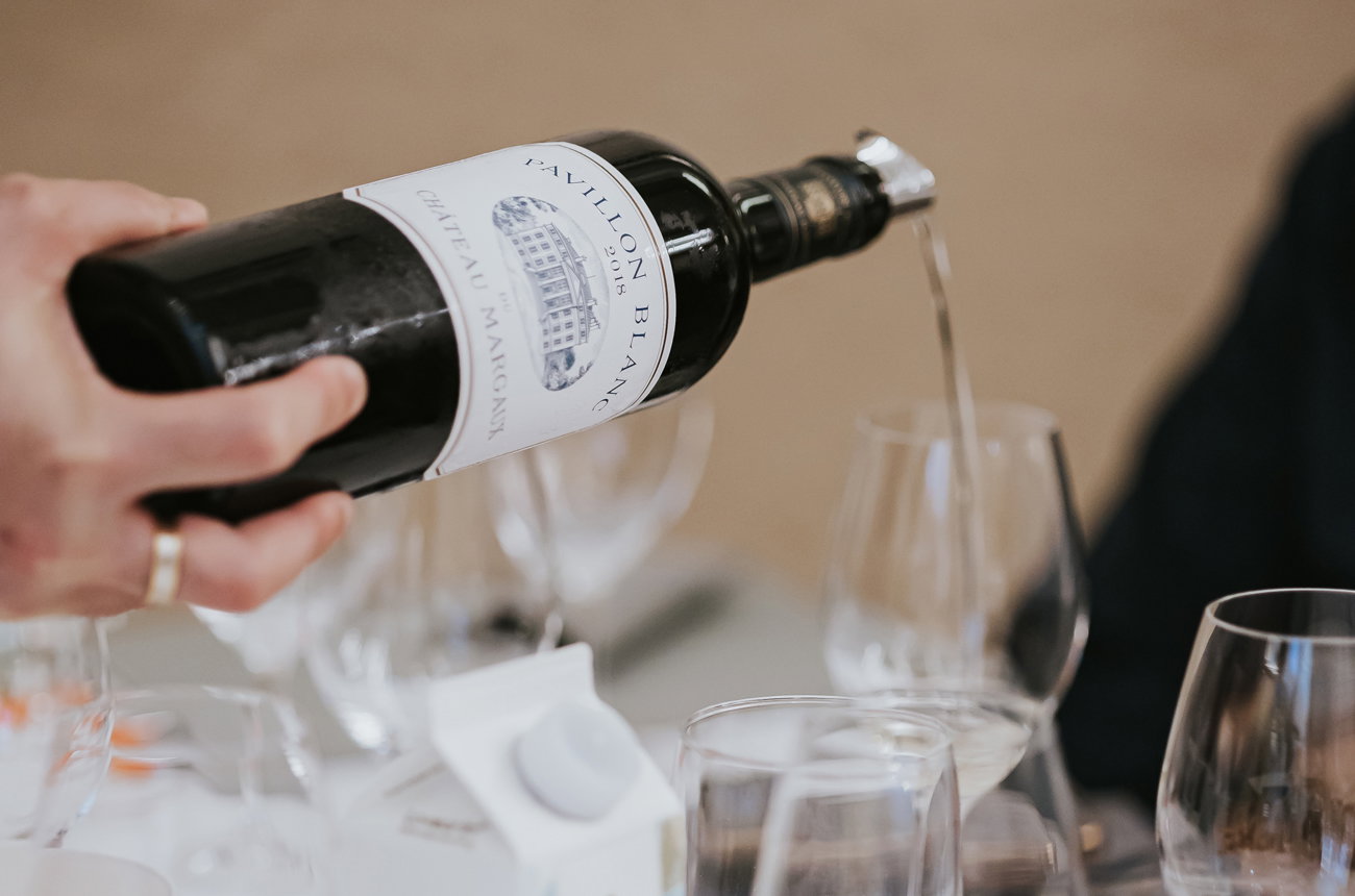 A person's hand holds a dark glass bottle with a white label pouring red wine into a clear glass on a table with other glassware.