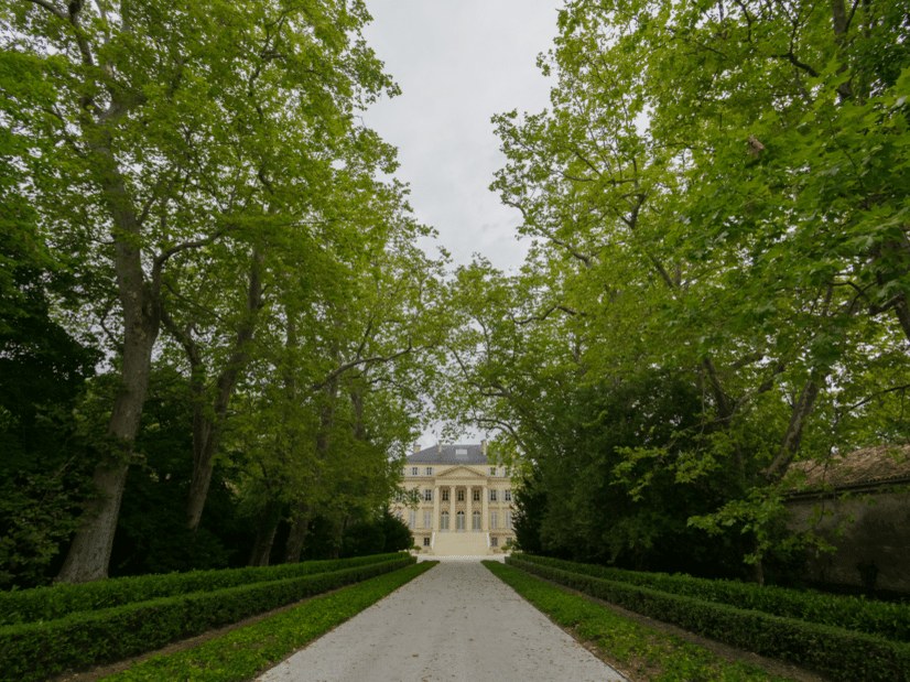 A long, tree-lined driveway leads to a grand, pale yellow Palladian mansion with columns and a dark roof under a cloudy sky.