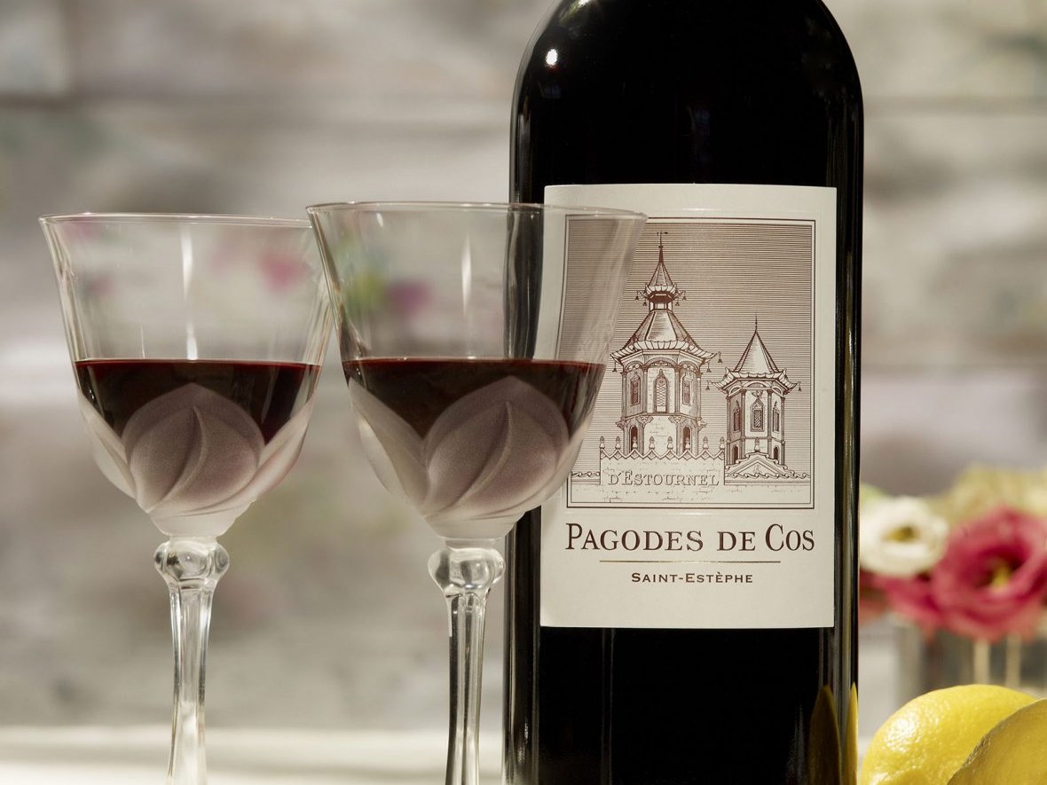 A dark bottle of Pagodes de Cos red wine stands next to two glasses filled with red wine, with cherries and lemons on a table.