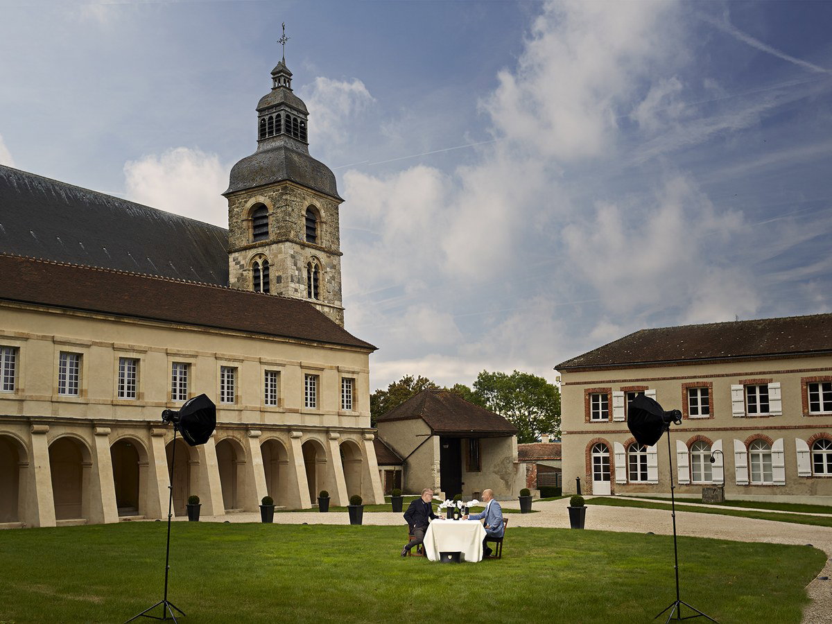 The exterior of the Abbey Saint Pierre d'Hautvillers features a stone bell tower, arcaded wings, and two men at a table on a lawn.