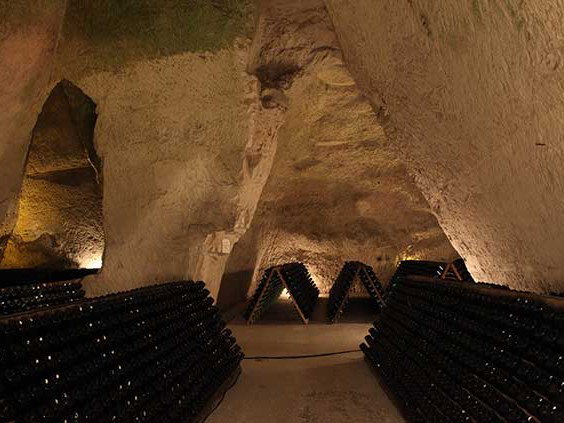 Underground chalk cellars with vaulted ceilings, illuminated to reveal rows of dark bottles stacked on wooden riddling racks.