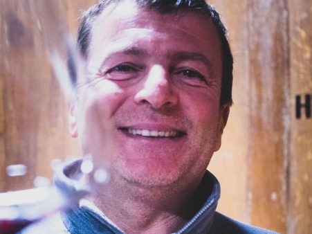 A smiling in a North Face fleece holds a glass of red wine in front of large wooden barrels labeled "HL 55".