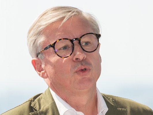 A man with glasses speaking, wearing a white shirt and green jacket