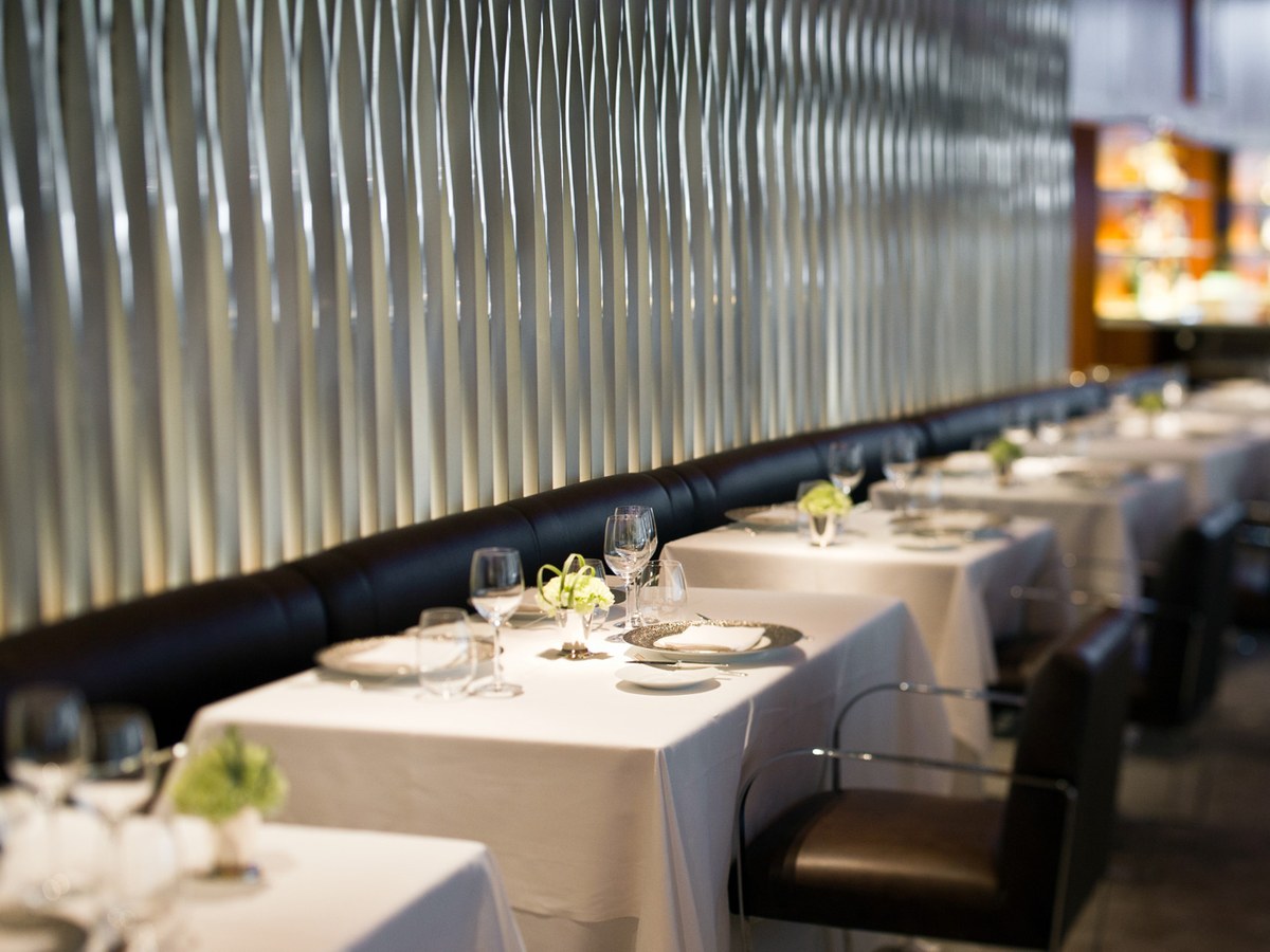 An elegant fine-dining restaurant interior with white-tablecloth tables, crystal glasses, and small floral centerpieces along a banquette.