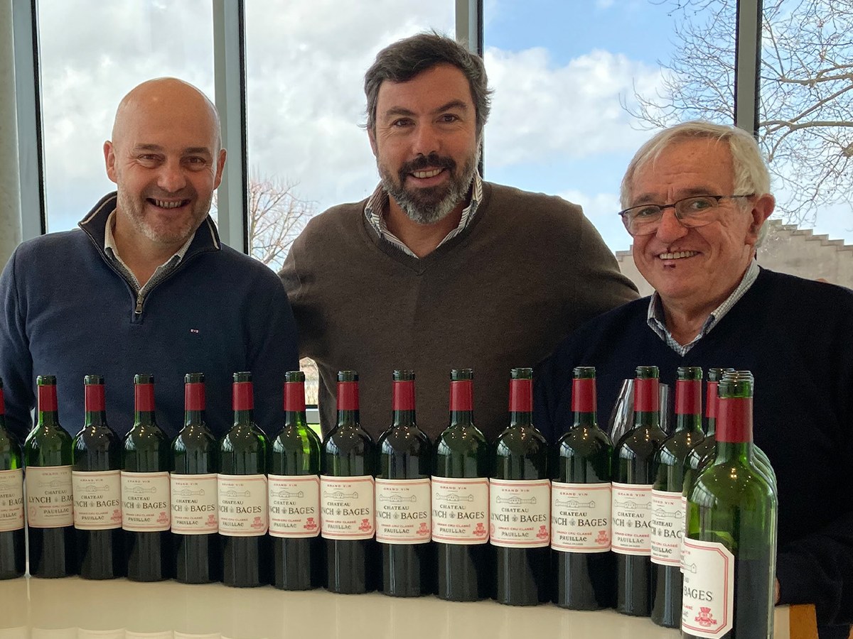 Three men stand behind bottles of Château Lynch-Bages in a winery.