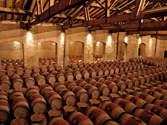 Hundreds of oak wine barrels are neatly stacked in rows inside a large, dimly lit cellar with stone walls and a high timber-beamed ceiling.