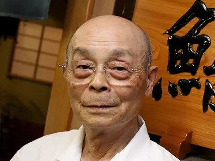 Jiro Ono, an elderly man with glasses and a white chef's coat, stands next to a wooden sign with black Japanese calligraphy.