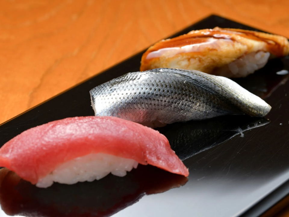 Three pieces of nigiri sushi, including tuna, silver-skinned fish, and cooked eel, are arranged on a black rectangular plate.