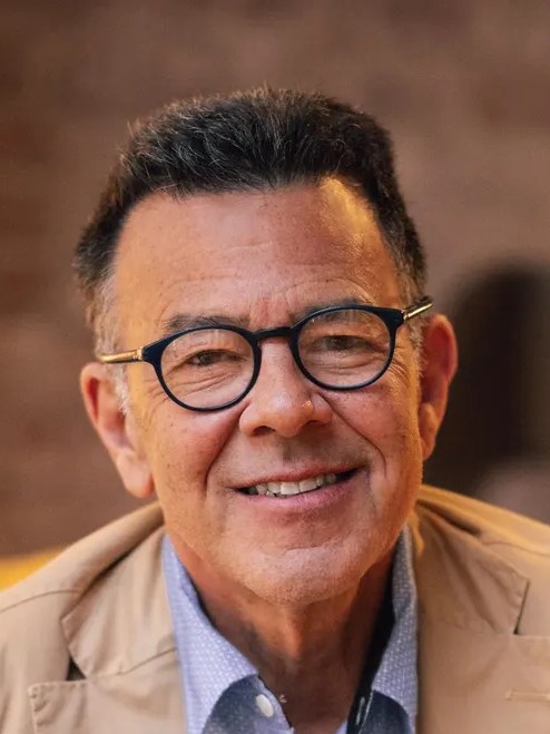 A smiling middle-aged man with dark hair and glasses, wearing a tan blazer over a light blue shirt, seated in a yellow chair.