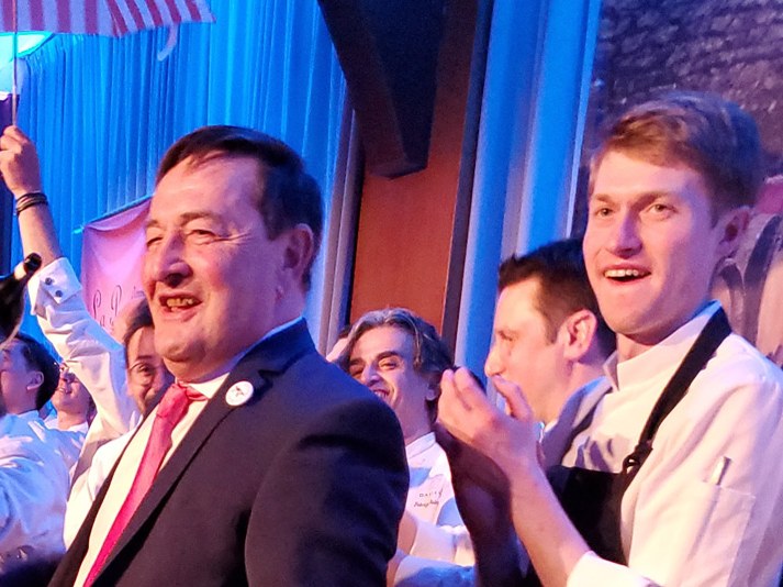 A celebratory crowd at La Paulée de New York, with people in chef whites and formal attire cheering, an American flag visible, and wine bottles on a