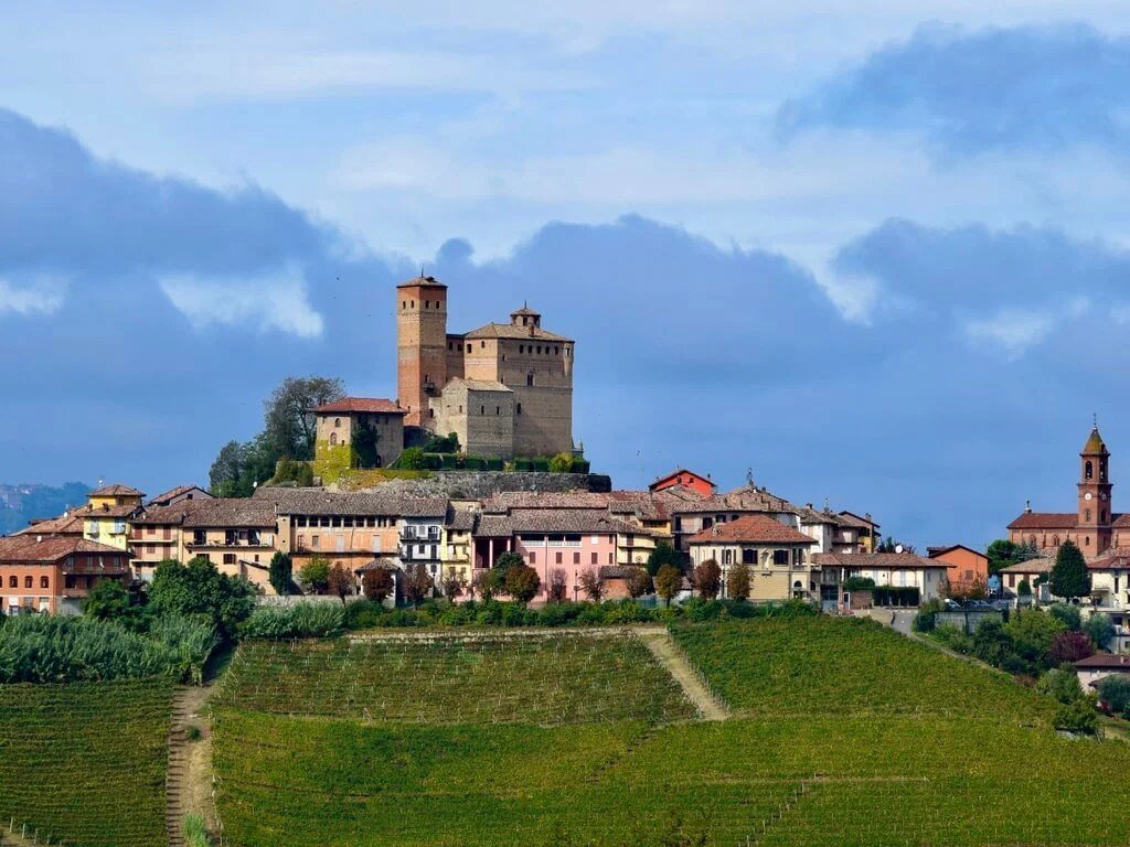 A medieval castle and village sit atop a hill, surrounded by green vineyards under a cloudy blue sky.