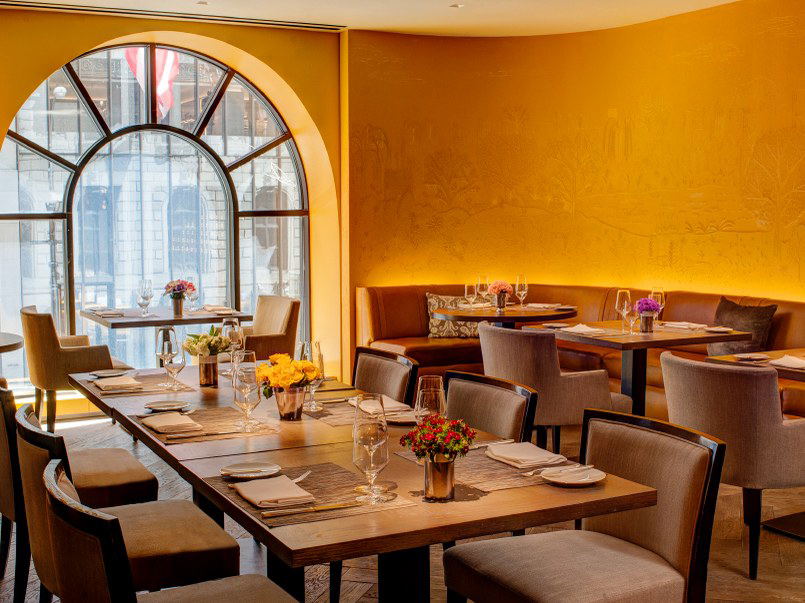An elegant restaurant dining room with golden walls, an arched window, and tables set with glassware and floral arrangements.
