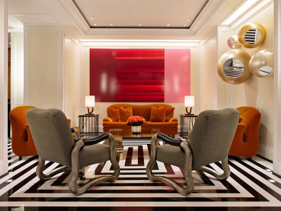 A luxurious hotel lounge with orange velvet sofas, sculptural gray armchairs, a red abstract artwork, and gold circular wall sculptures.