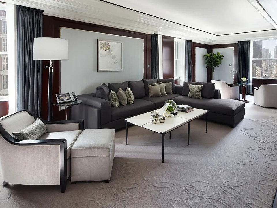 A luxurious hotel suite living room with a dark sectional sofa, light armchairs, patterned carpet, floor lamps, and city views.