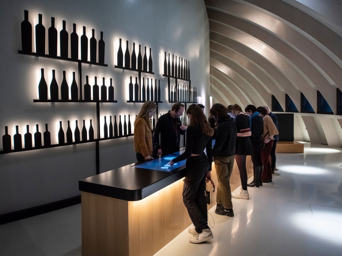 Visitors interact with a large touchscreen display at a tasting bar, with backlit wine bottle silhouettes on the wall.